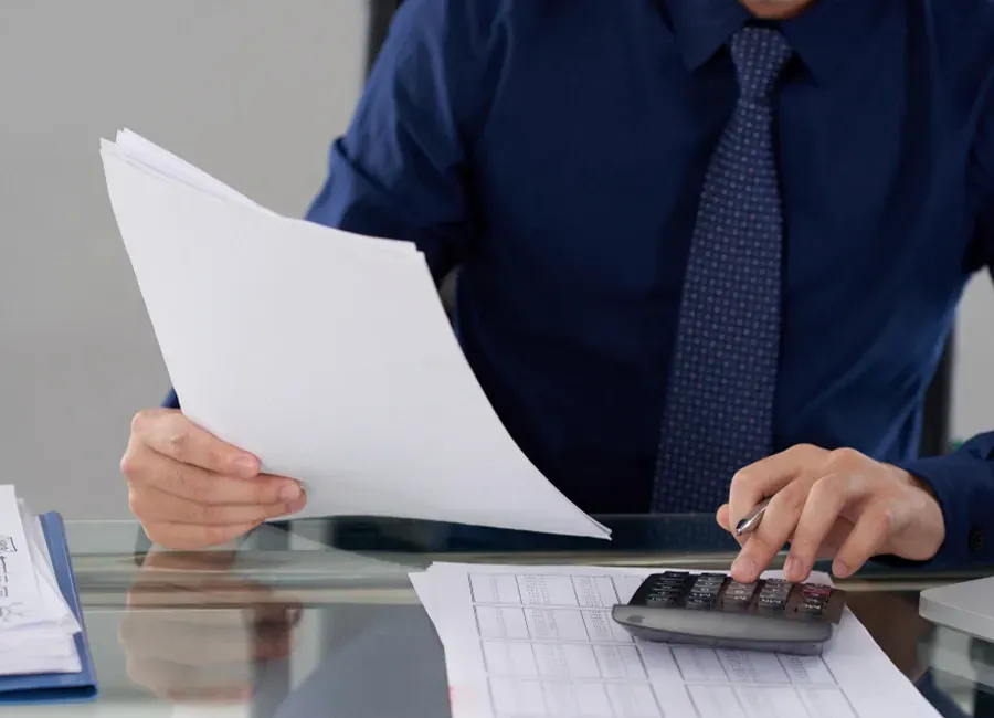 Man in blue shirt and tie using a calculator, holding papers, possibly doing calculations in an office setting.