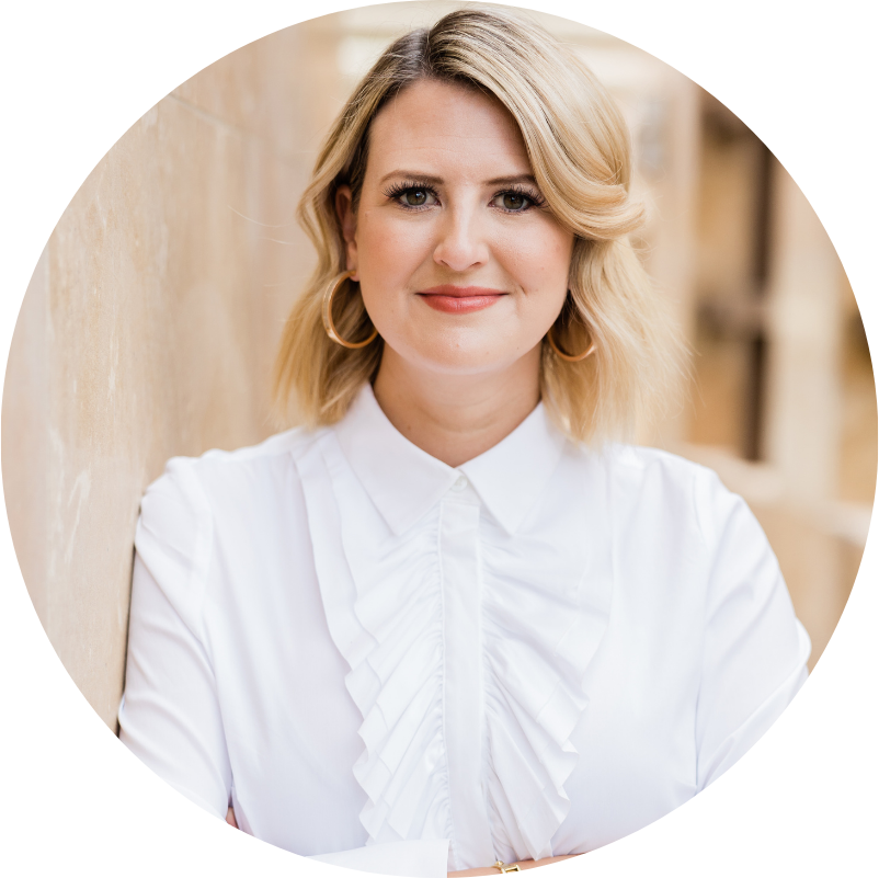 Woman with blonde hair, in a white ruffled shirt, smiles, leaning against a wall, wearing gold hoop earrings.