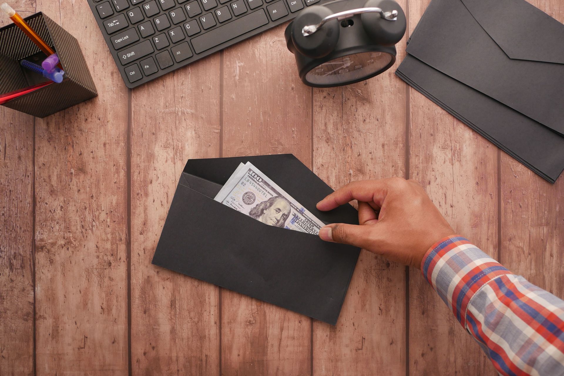 Hand placing cash into a black envelope on a wooden desk with office items nearby