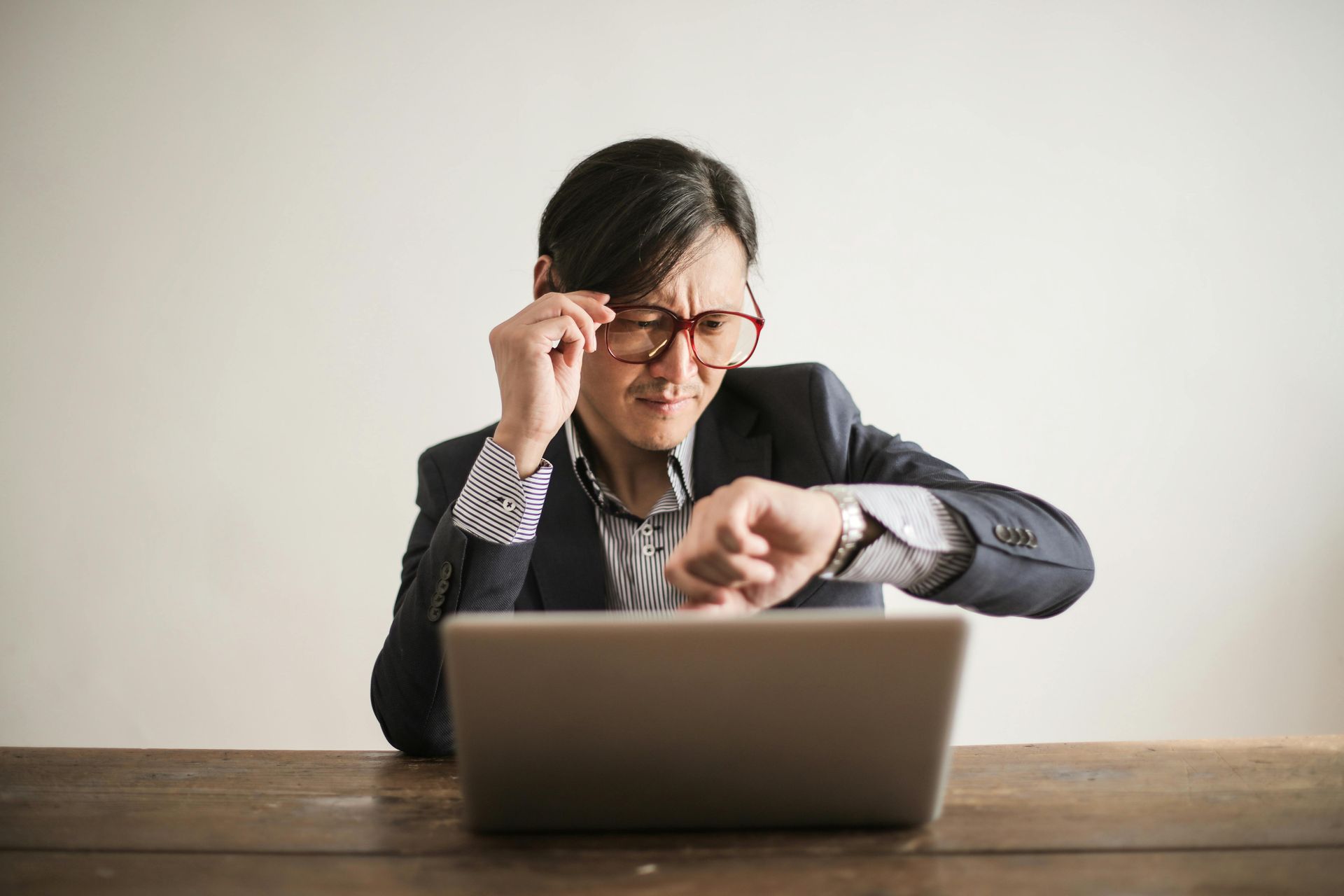 A man is sitting at a desk looking at his watch while using a laptop computer.