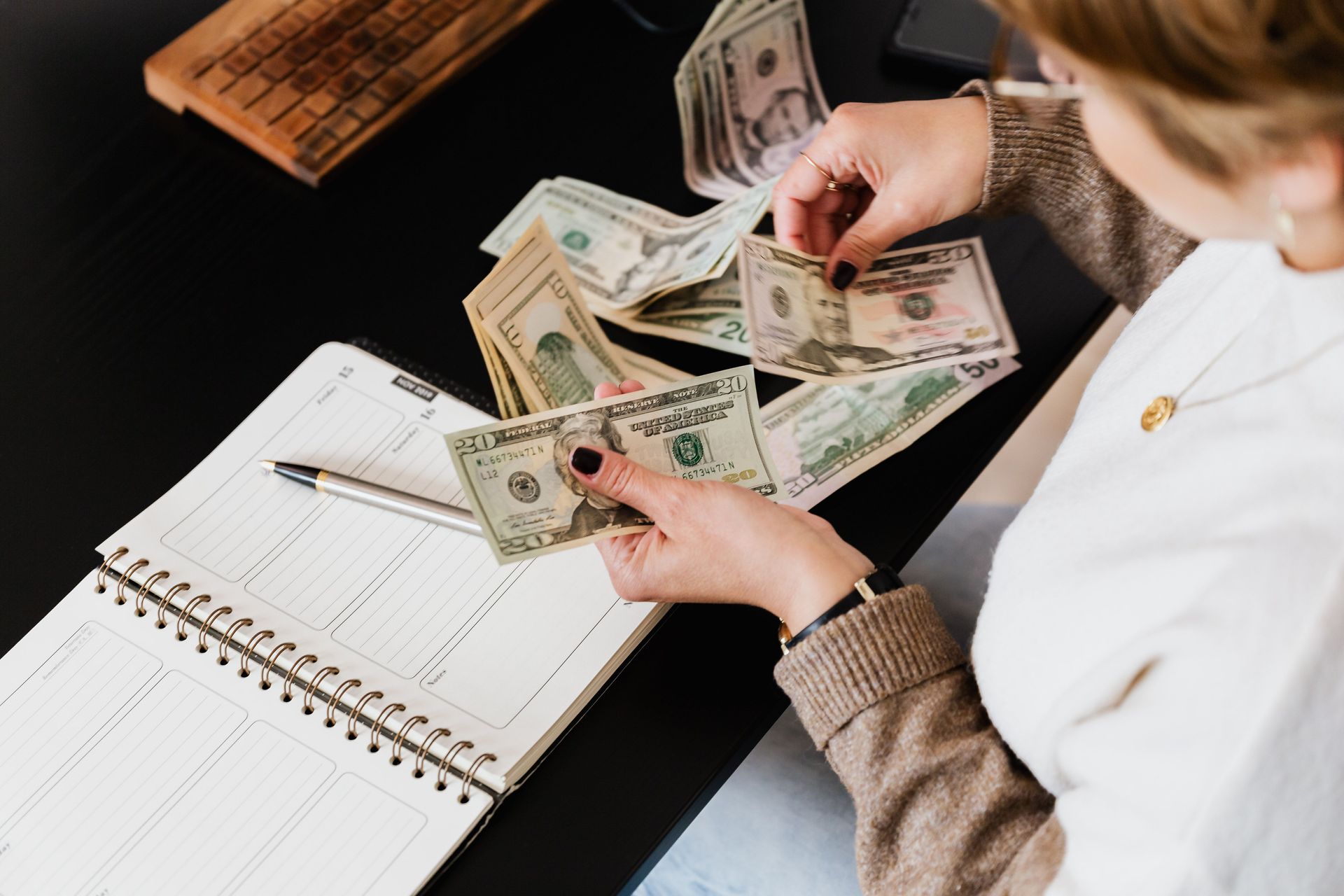 Person counting U.S. dollar bills beside an open notebook and ledger on a desk