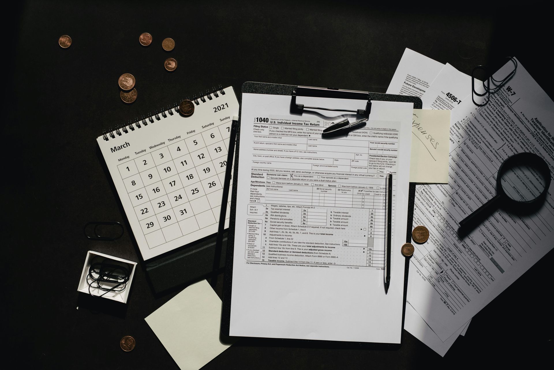 Desk with scattered papers, a clipboard, magnifying glass, notebook, and coins on a dark background.