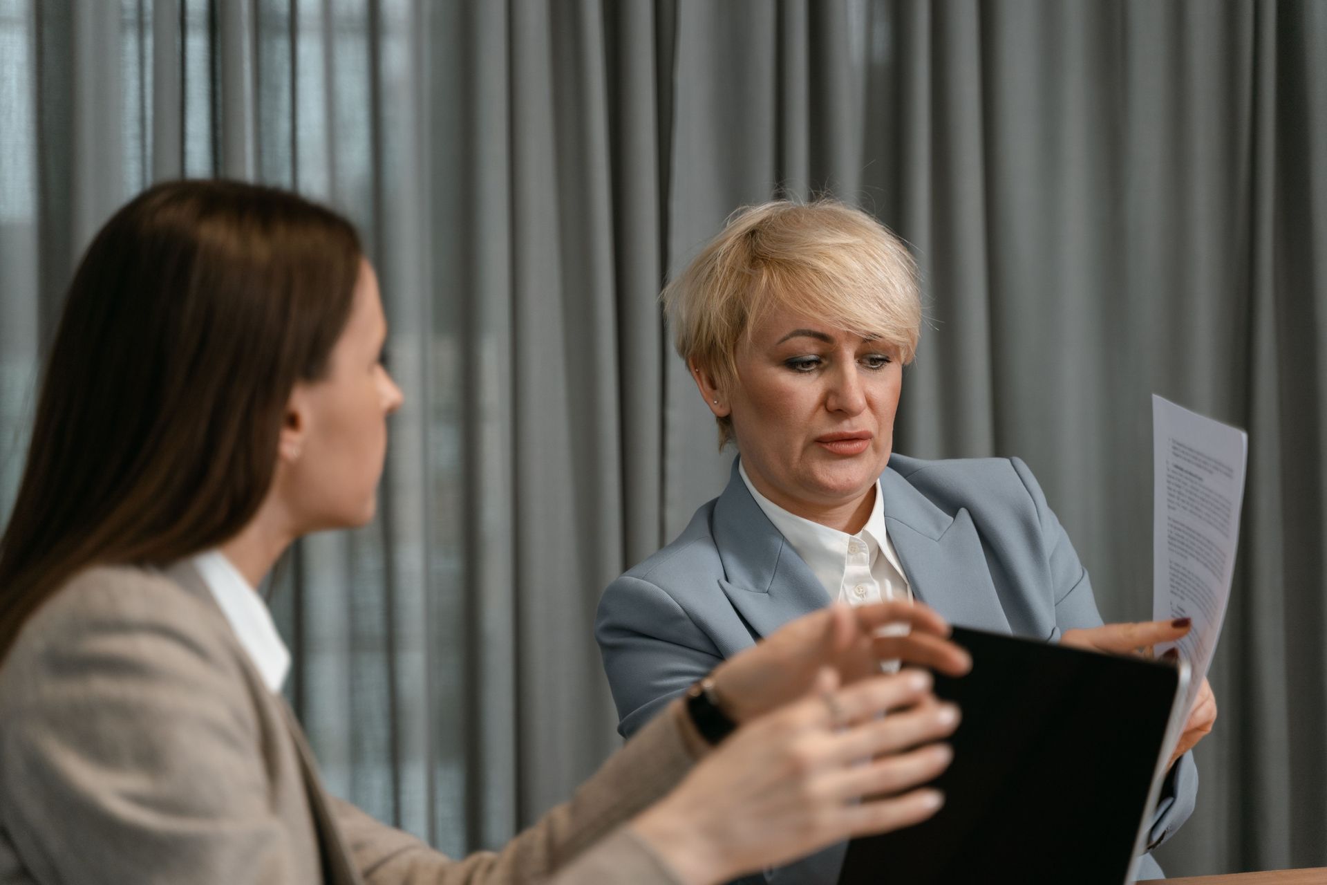 Two people in a meeting, one holding papers and the other gesturing beside a laptop in an office.