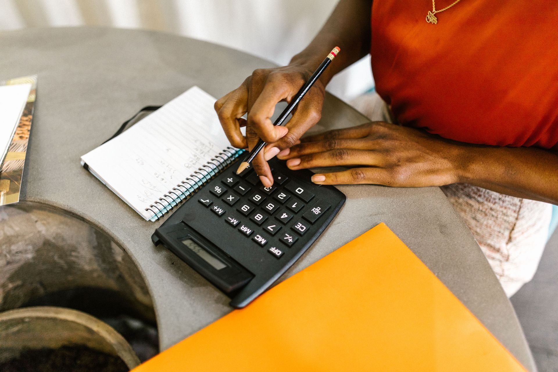 Hands using a calculator and pen beside a notebook on a table with an orange folder