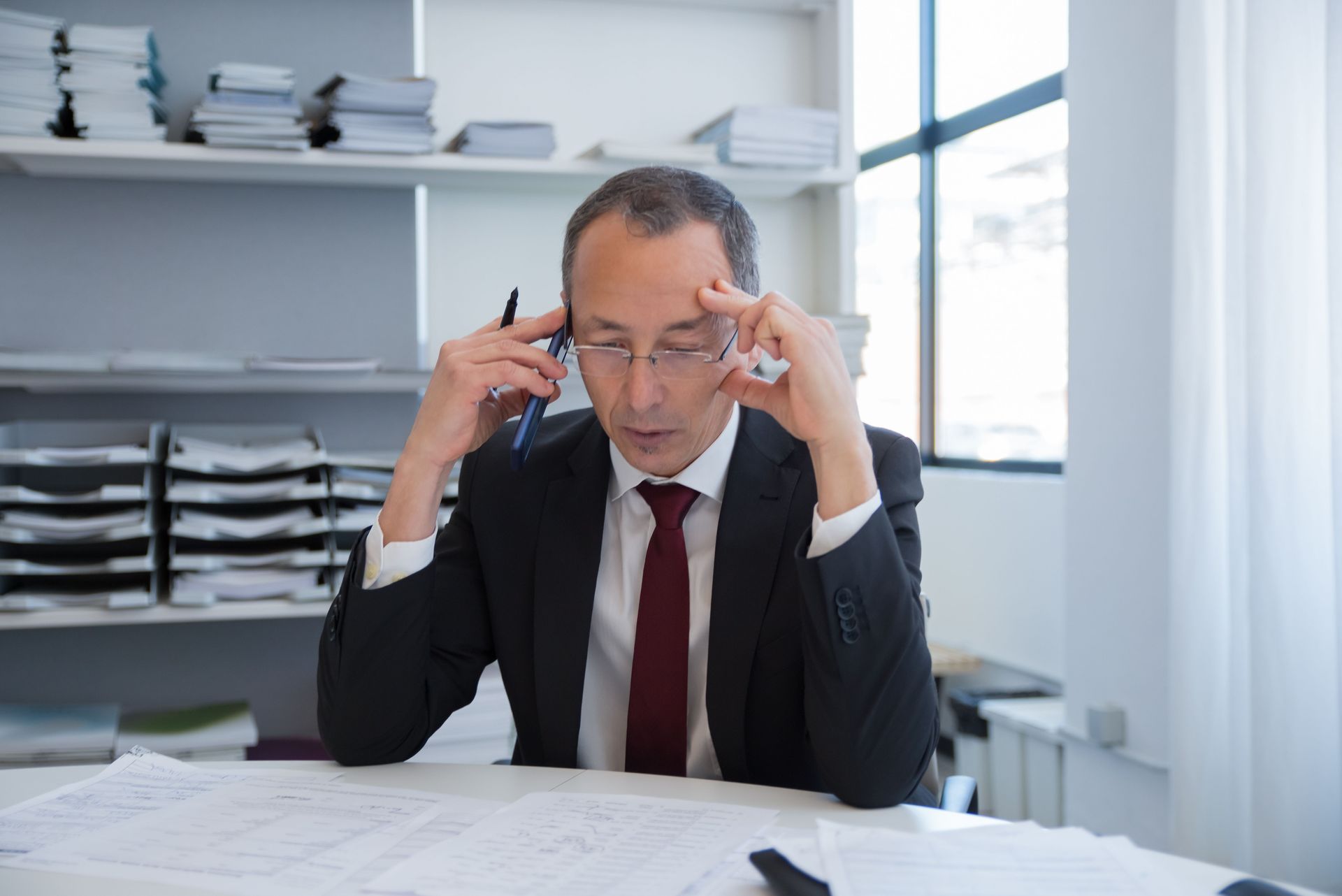 Office worker in a suit rubbing his temples at a desk with papers and shelves of files behind him