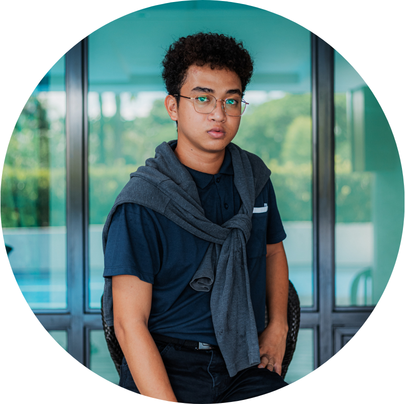 Young man with glasses, sweater around shoulders, sitting outdoors in front of a blue glass wall.