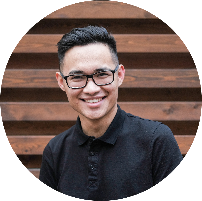 Smiling Asian man with glasses in a black polo shirt stands in front of wooden slats.