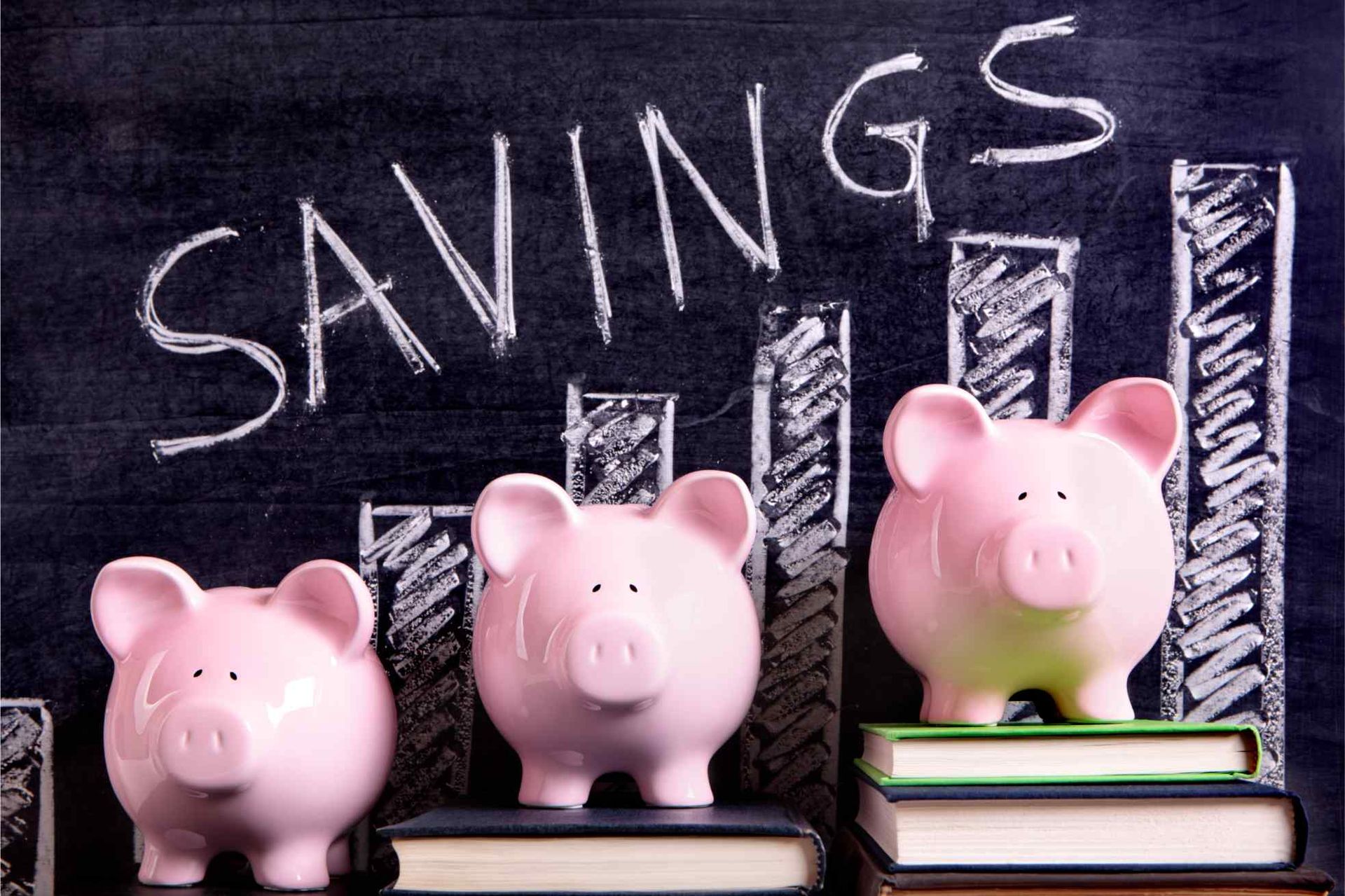 Three pink piggy banks are sitting on top of books in front of a blackboard that says savings
