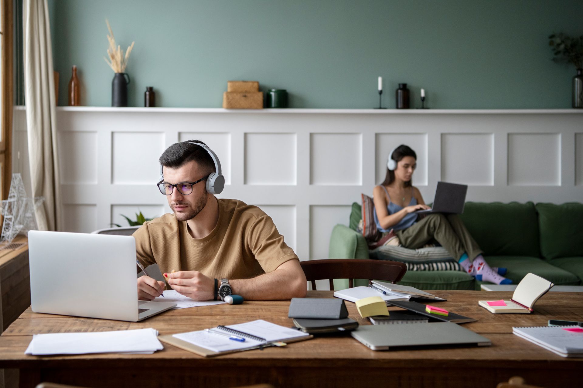 A man is sitting at a table using a laptop while a woman sits on a couch.