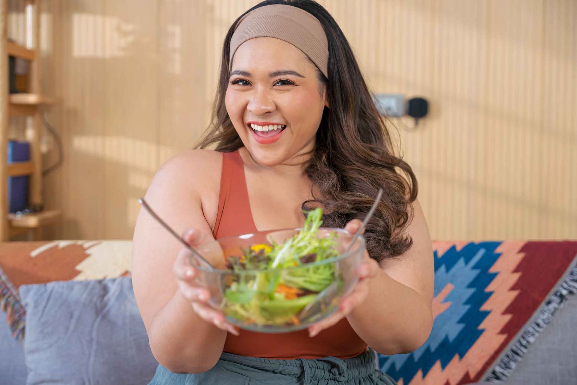 A woman is sitting on a couch holding a bowl of salad.