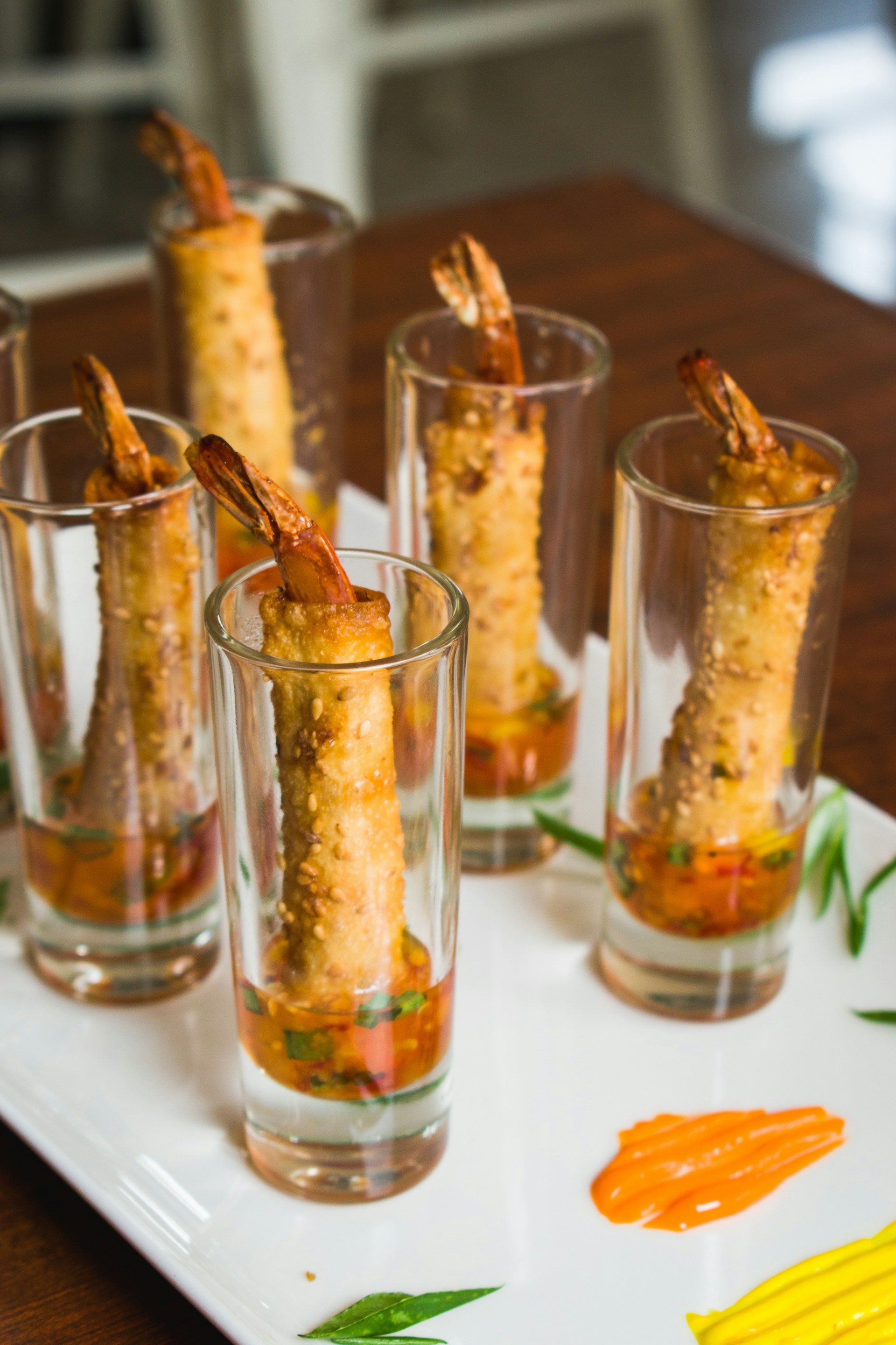 Shrimp skewers in tall glasses on a white tray, with orange sauce and garnish