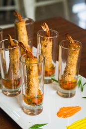 Shrimp skewers in tall glasses on a white tray, with orange sauce and garnish