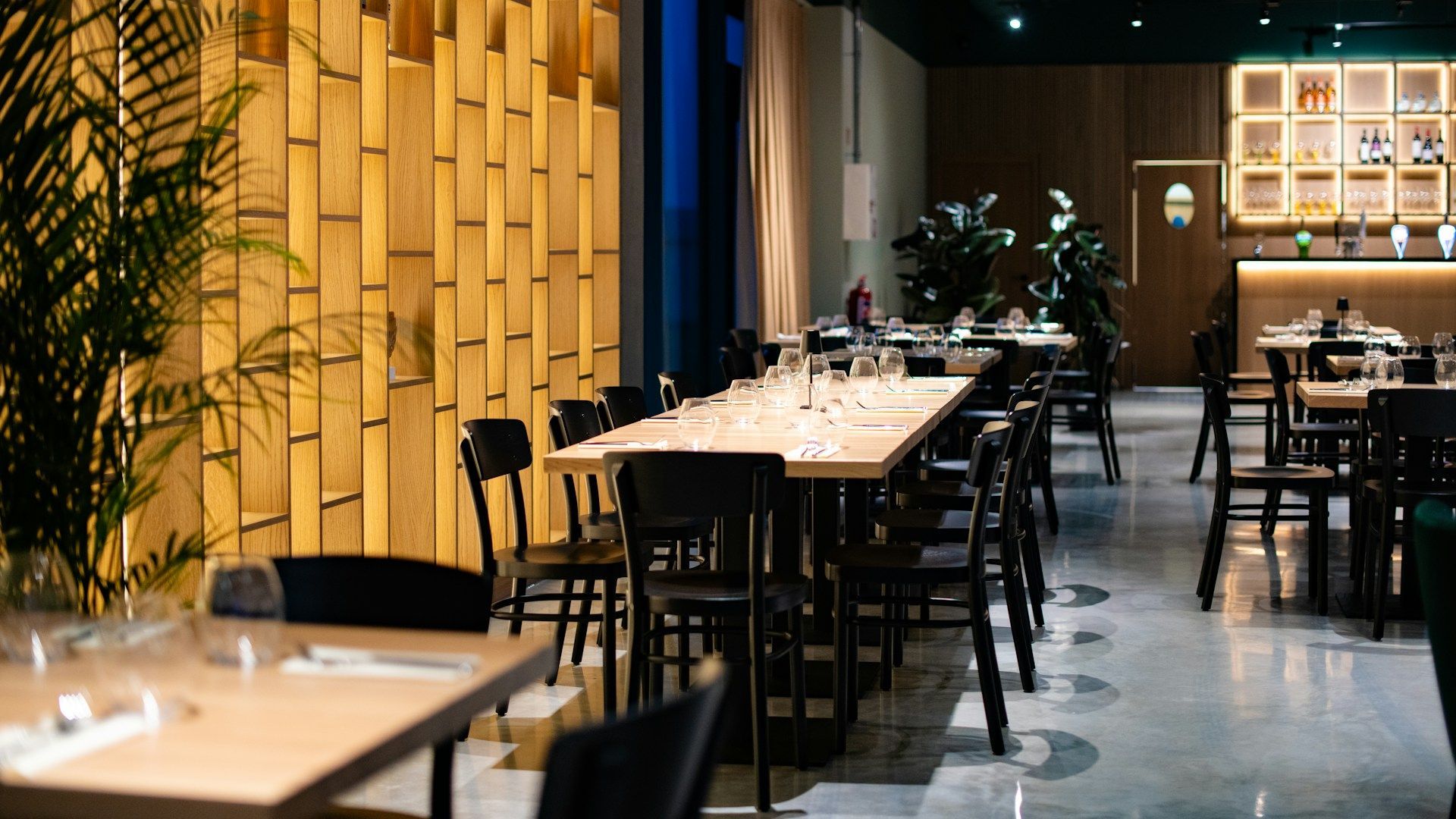 Modern restaurant interior with empty wooden tables, black chairs, warm lighting, and a glossy floor.
