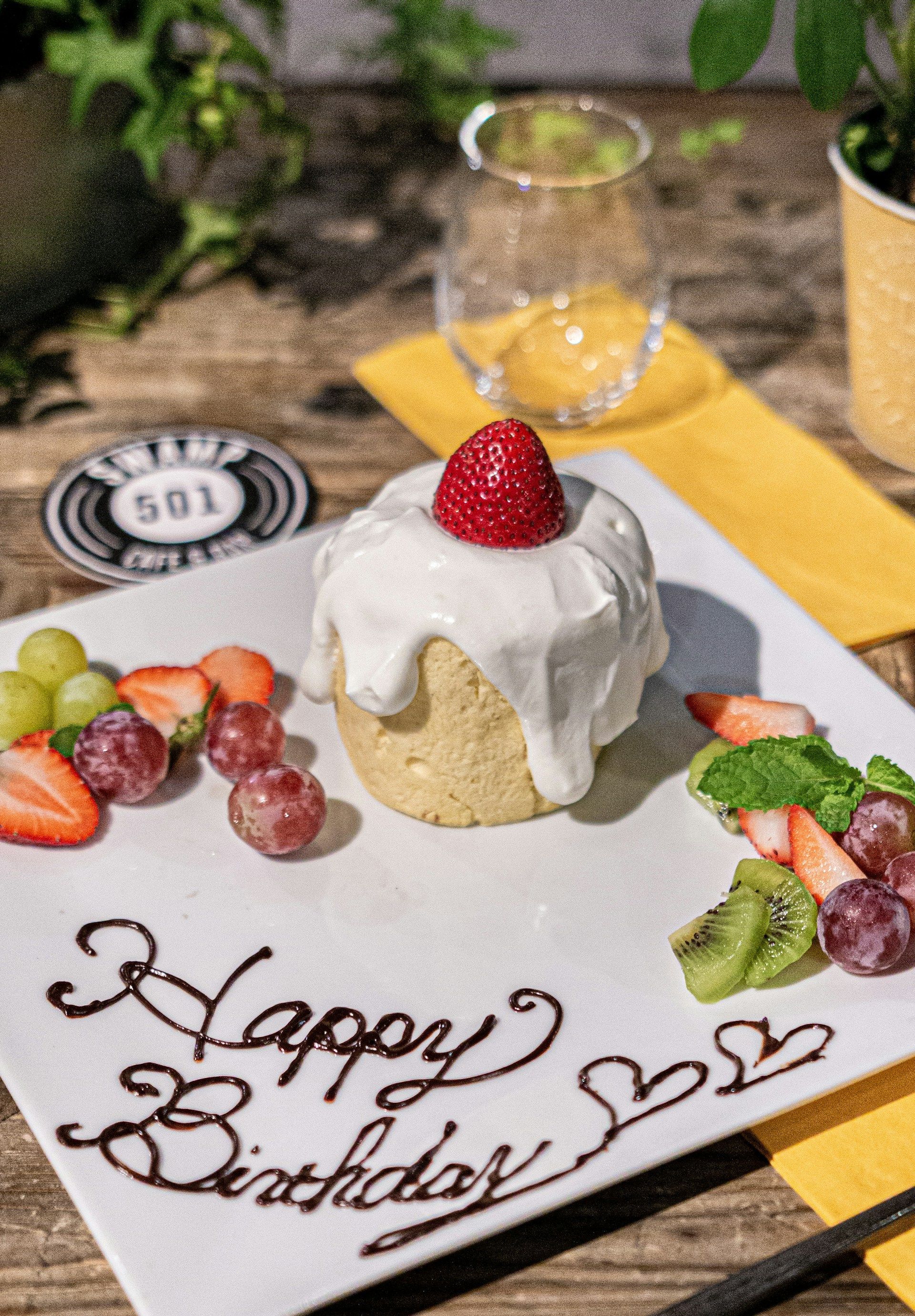 Birthday dessert with strawberry topping, “Happy Birthday” writing, and colorful fruit on a table