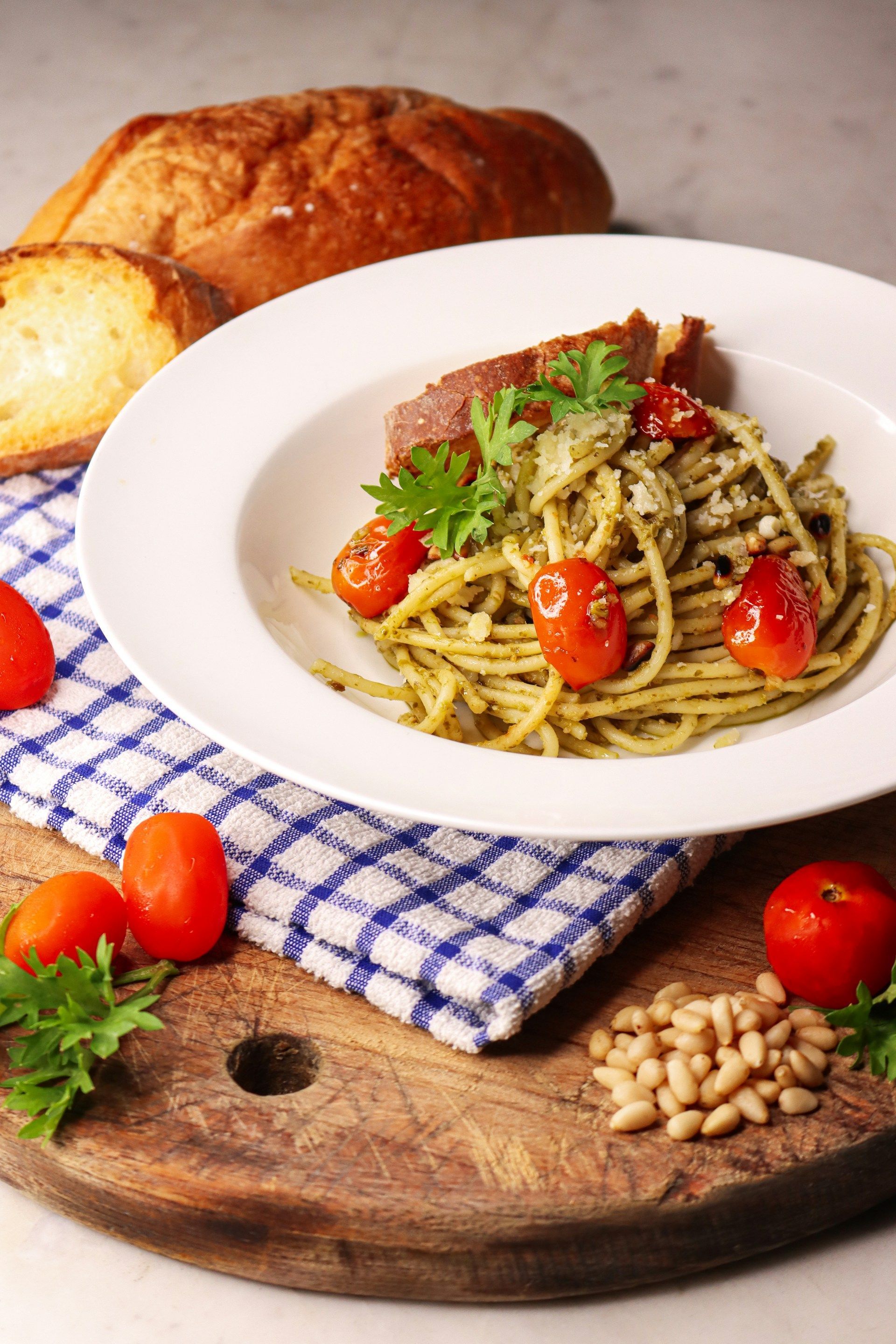 Pasta with pesto and cherry tomatoes in a white bowl, with bread, pine nuts, and parsley on a wooden board