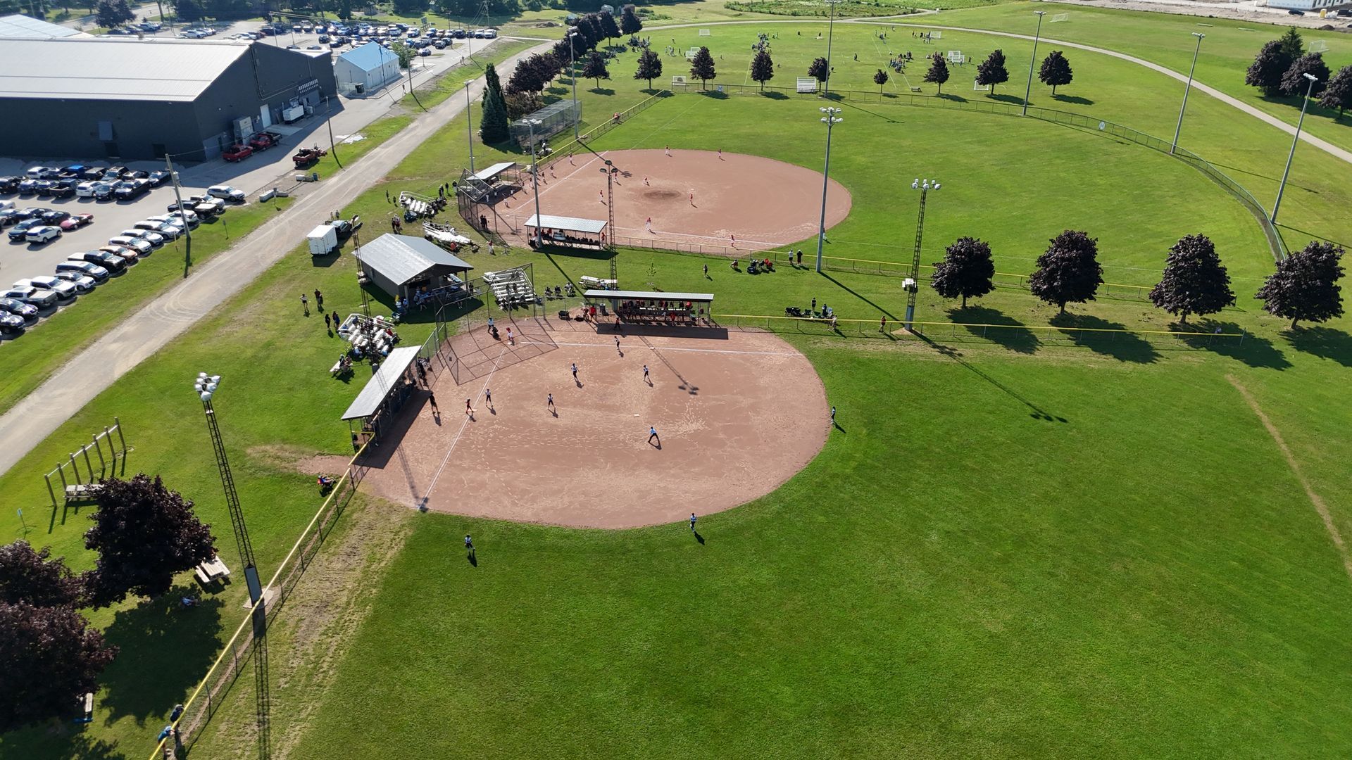 Aerial view of two baseball fields, one with a game in progress, surrounded by green grass and trees.