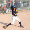 A child in a baseball uniform swings a bat at a ball during a game on a dirt field.
