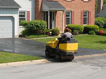 A man is driving a yellow roller on a driveway in front of a house.