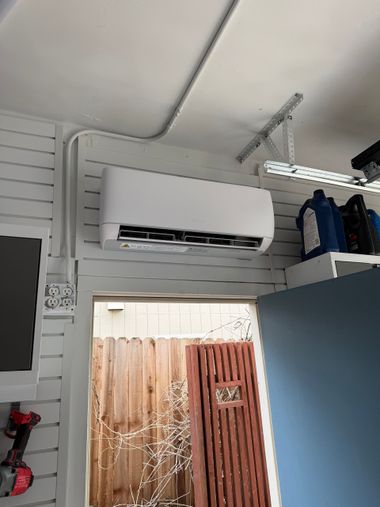 White wall-mounted air conditioner above a doorway in a small room with slatted walls and shelves.