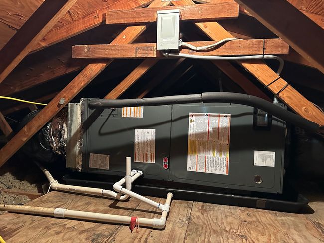 Black HVAC unit in an attic with exposed wooden beams and white piping.