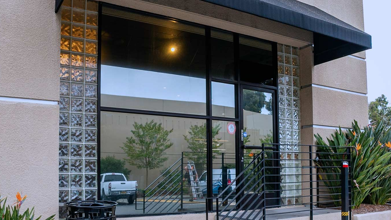 Exterior view of a building with a large glass window and black awning. There's a truck reflected in the glass.