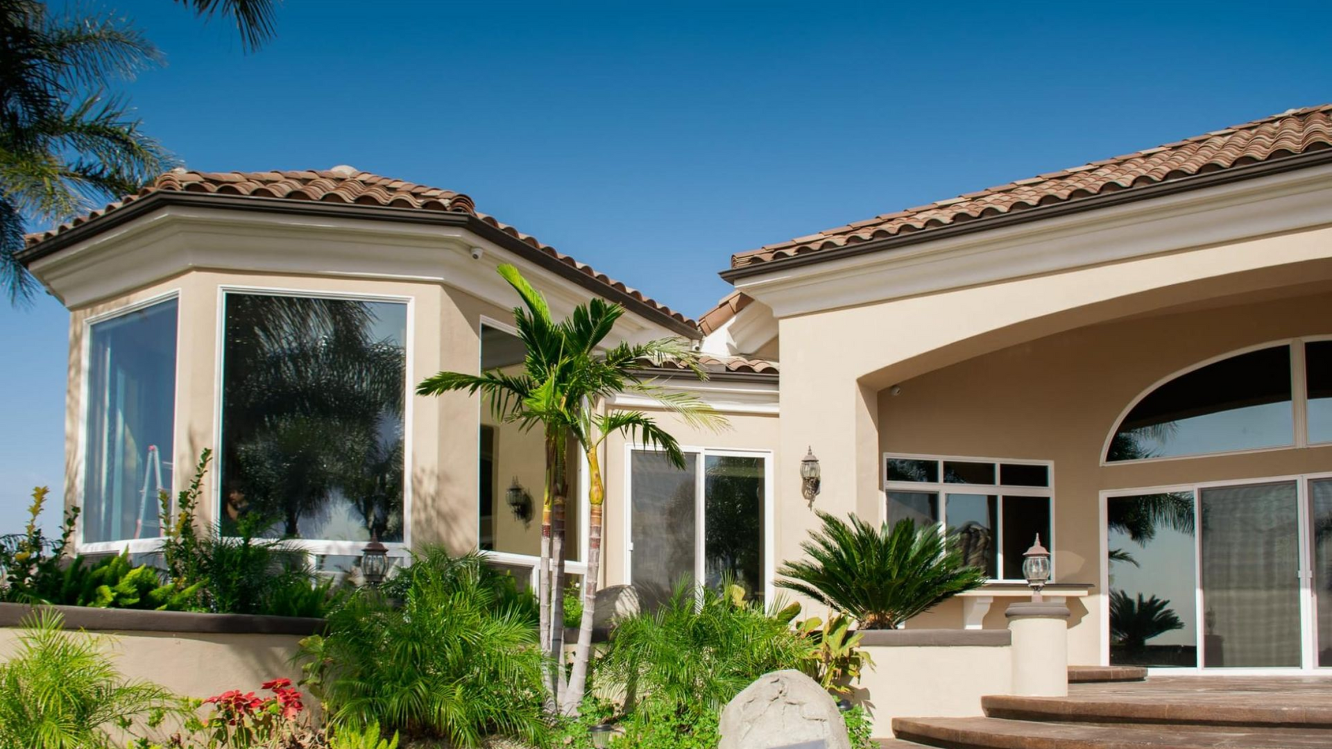 Mediterranean-style home with stucco walls, tile roof, large windows, and lush landscaping under a blue sky.