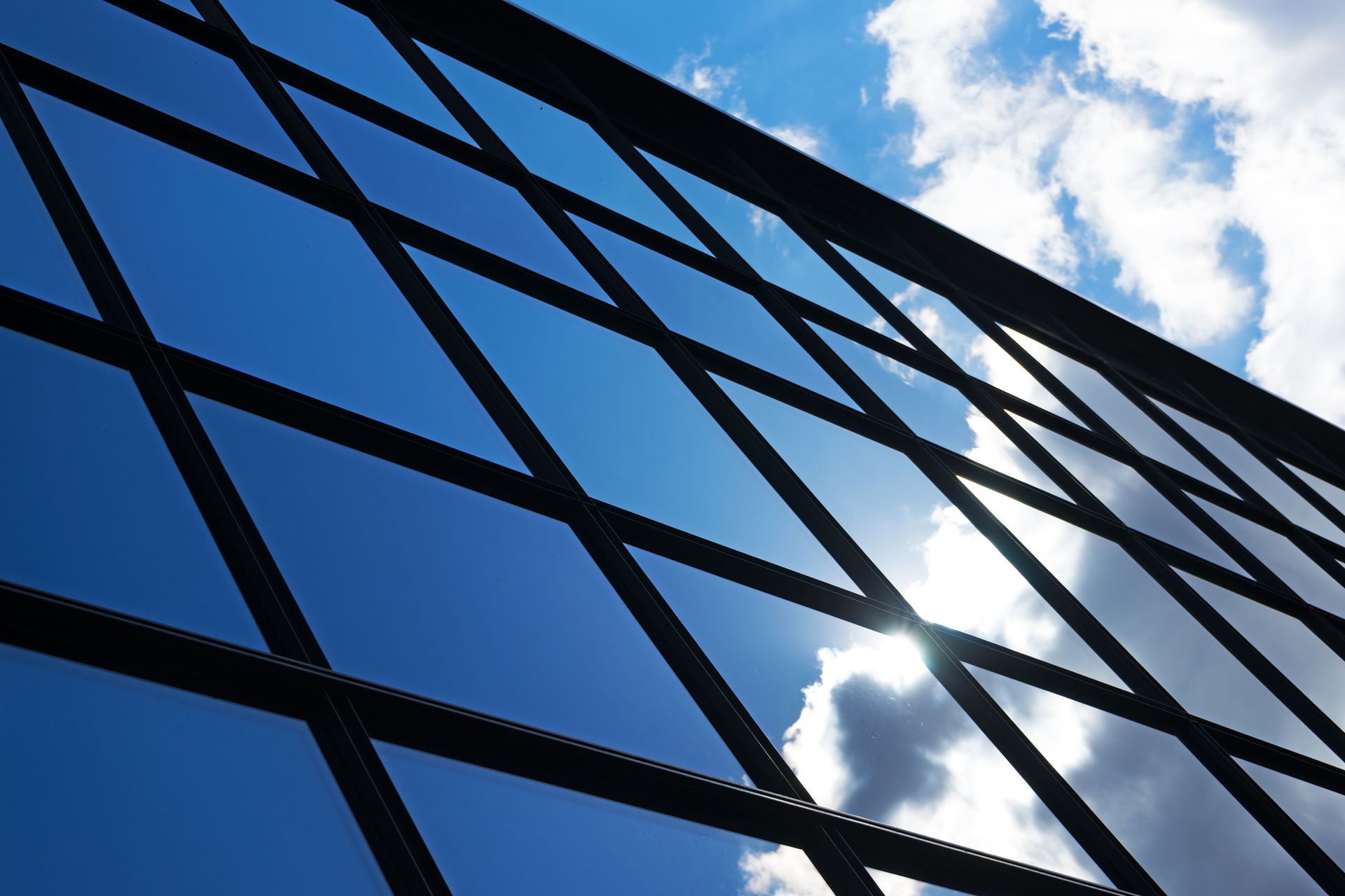 Diamond pattern black metal fence against a bright blue sky with clouds and sun.