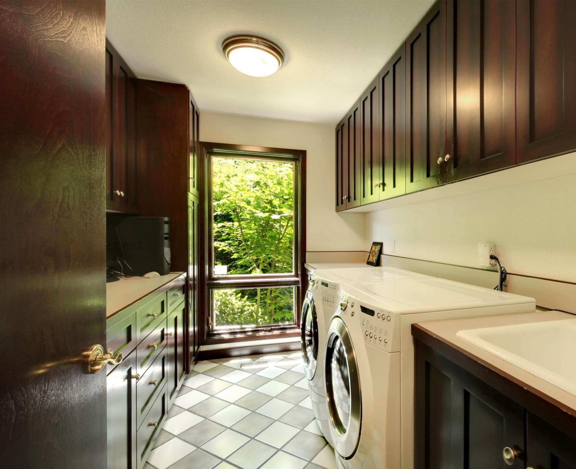 Narrow laundry room with dark cabinets, white appliances, tiled floor, and an open door to greenery.