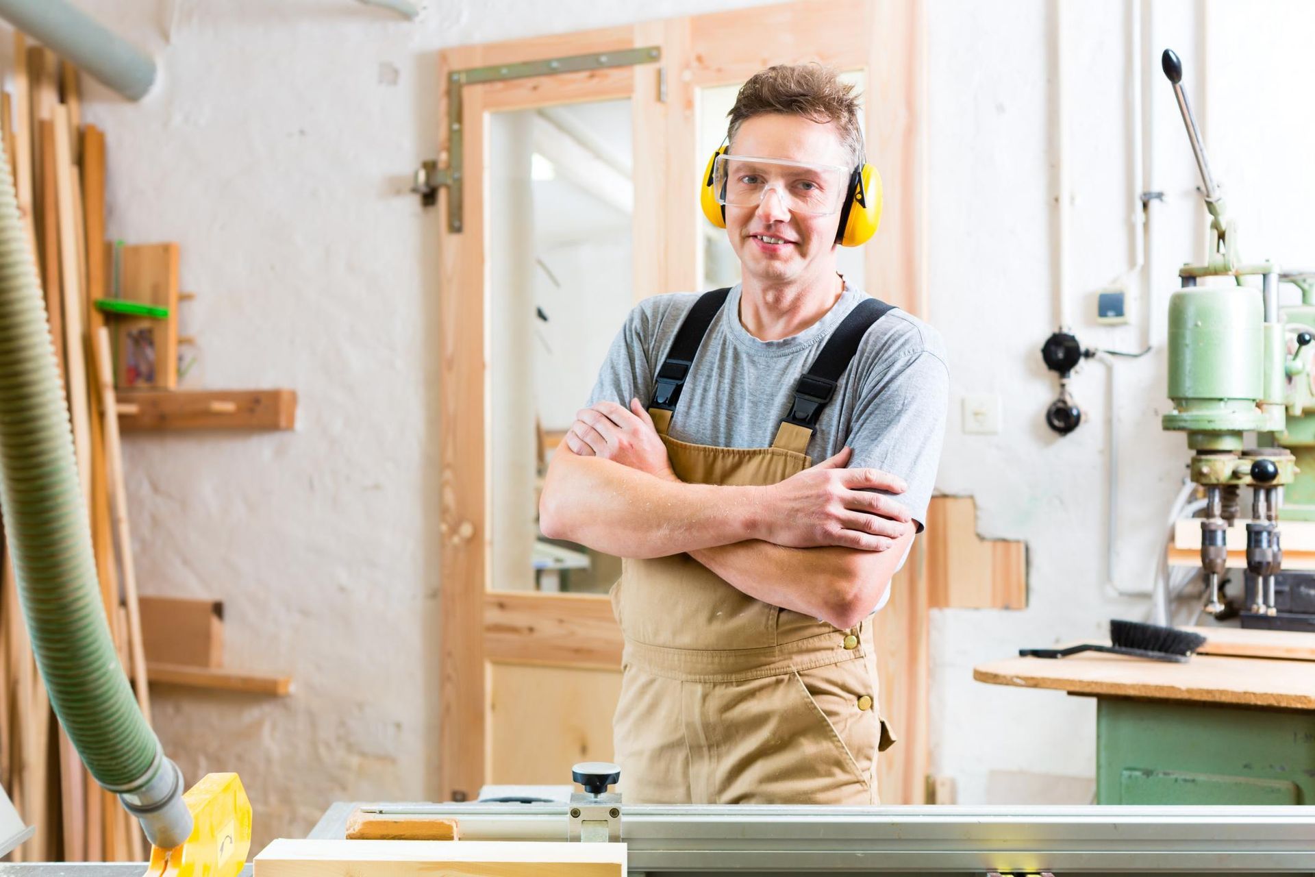 Woodworker in a workshop, smiling with arms crossed beside a workbench and drill press.