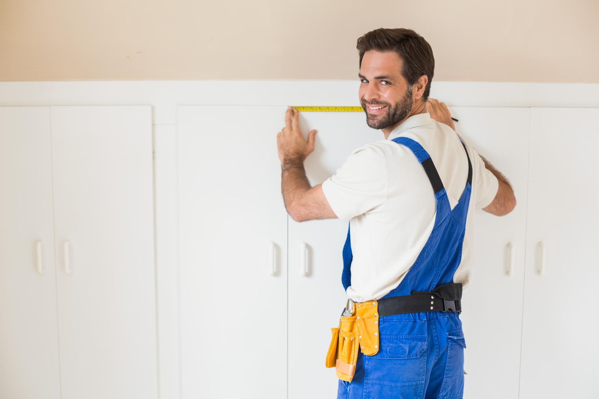 Contractor holding a tape measure against white cabinets, wearing blue overalls and a tool belt