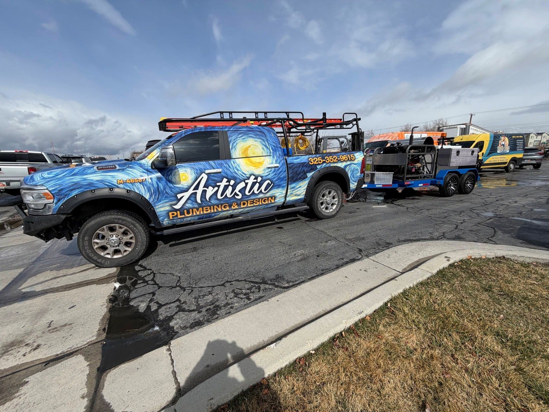 Blue “Artistic” tow truck with a trailer parked on a cracked road under a cloudy sky.