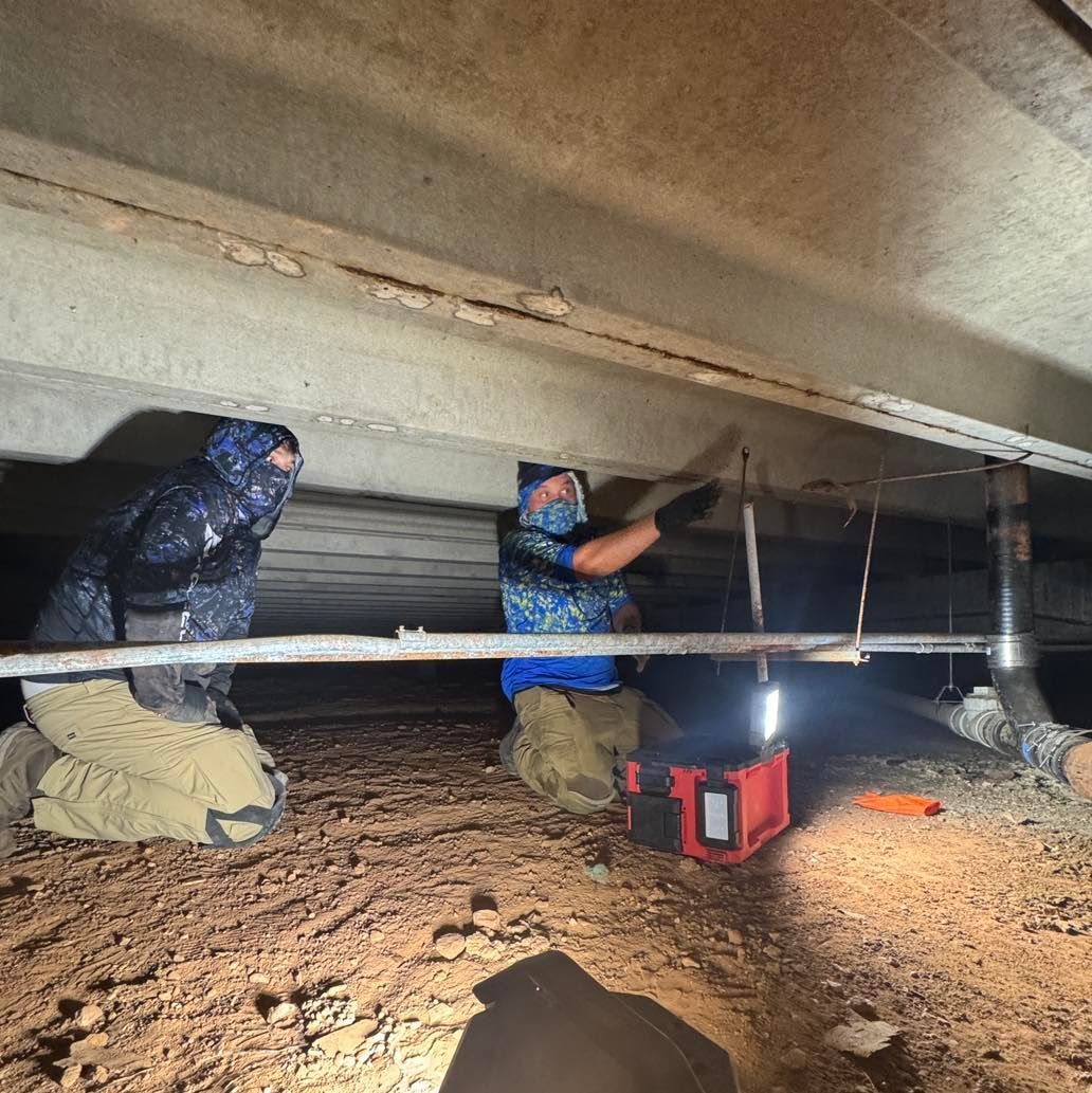 Workers in a crawl space install a beam under a low ceiling, lit by a red work light.
