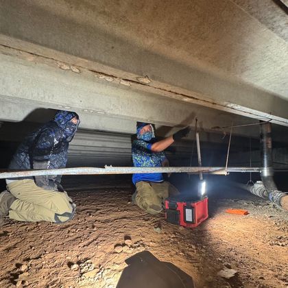 Workers in a crawl space install a beam under a low ceiling, lit by a red work light.
