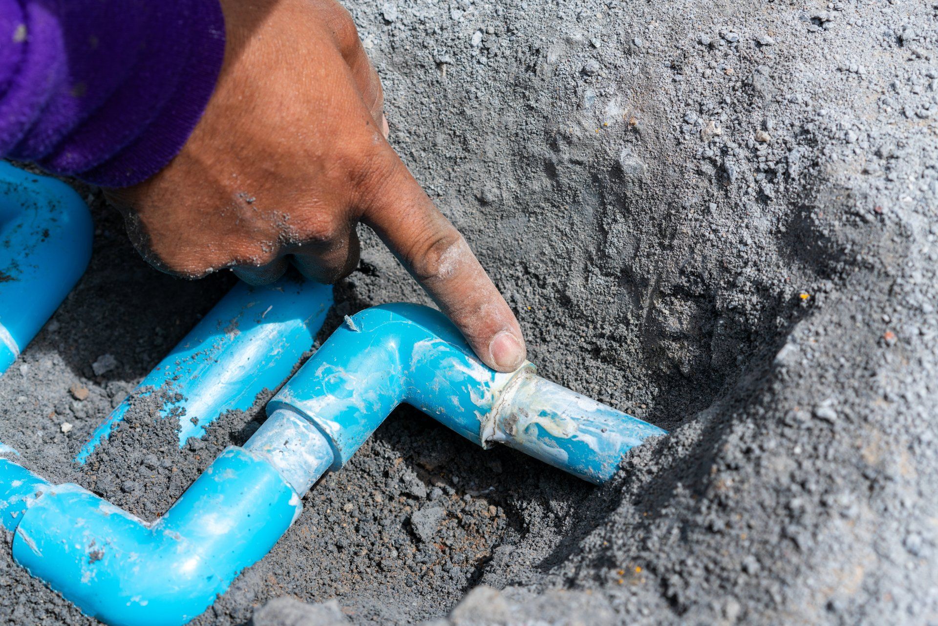 Hand placing a blue plastic pipe fitting into wet cement in a trench