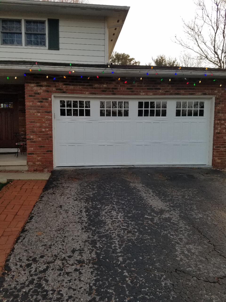 White two-car garage with holiday lights and a dark driveway in front of a brick house