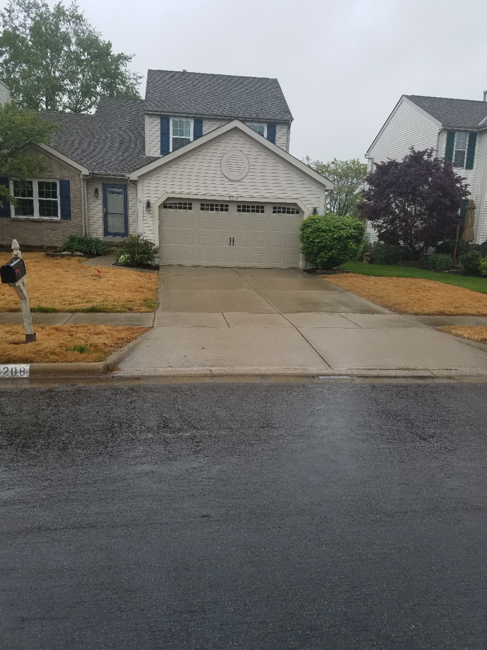 Suburban house with beige garage, wet driveway, and landscaped front yard on a rainy day