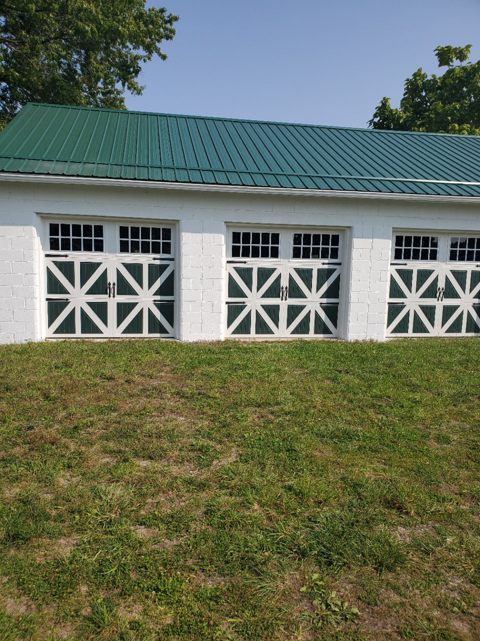 White barn with green metal roof and three white-trimmed doors on a grassy slope under a blue sky