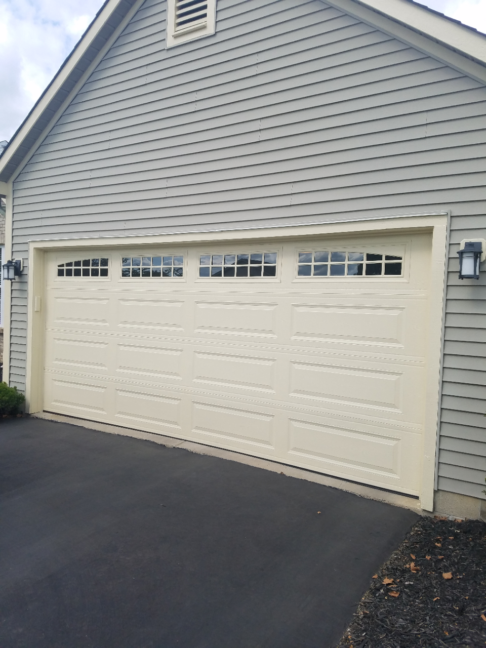 Cream-colored two-car garage with decorative windows on a gray-sided house, beside a paved driveway