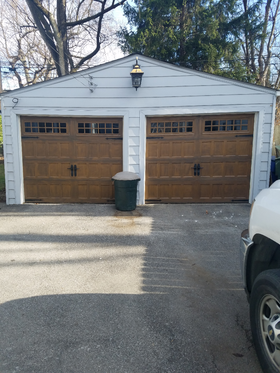 White two-car garage with brown doors, a trash bin, and a vehicle edge in the driveway.