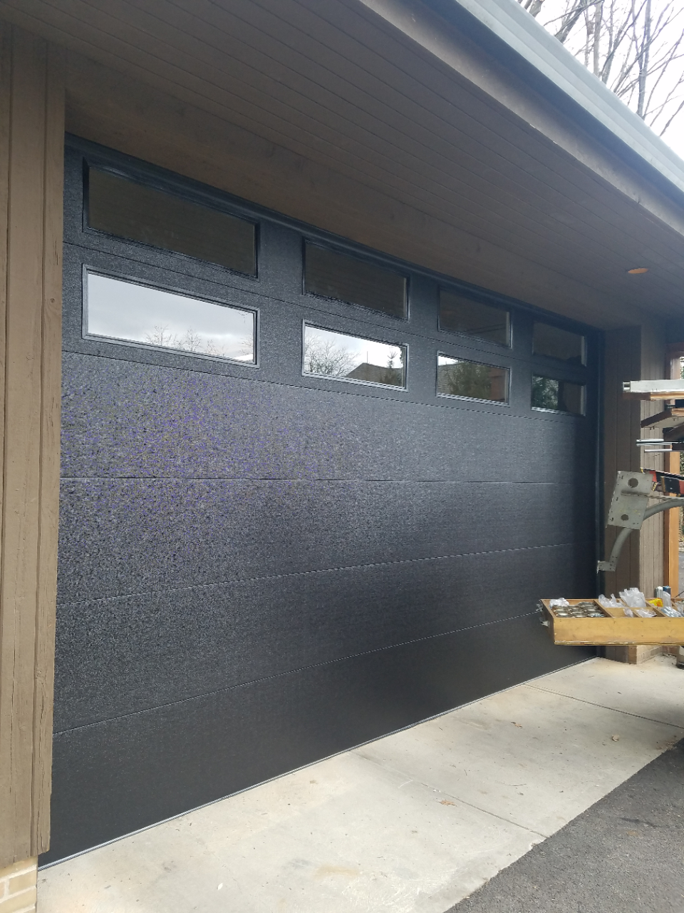 Black garage door with small windows on a driveway, partially open, with wood siding and roof overhang.
