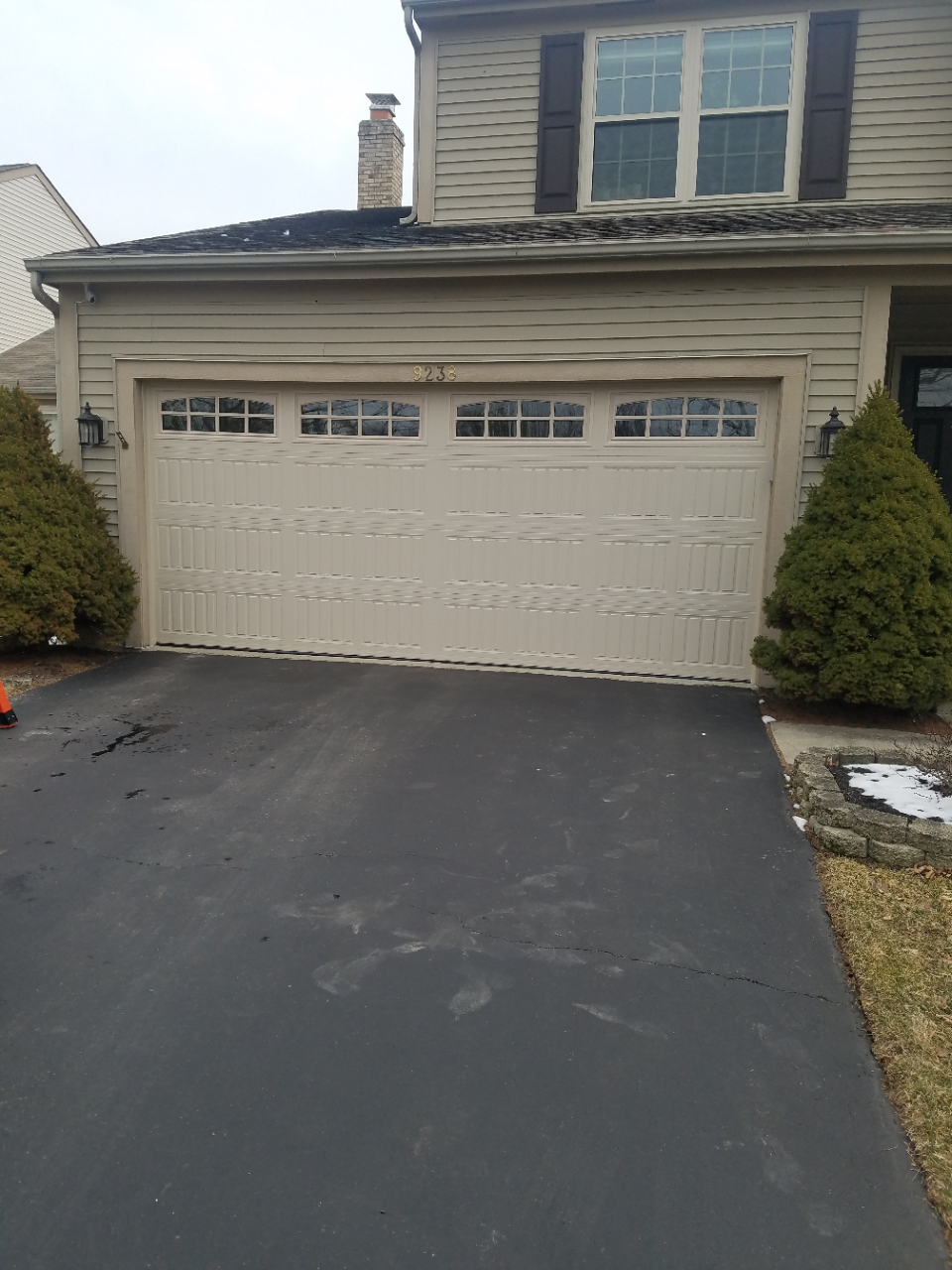 Two-story suburban house with a beige garage and dark driveway, flanked by trimmed shrubs and light snow patches