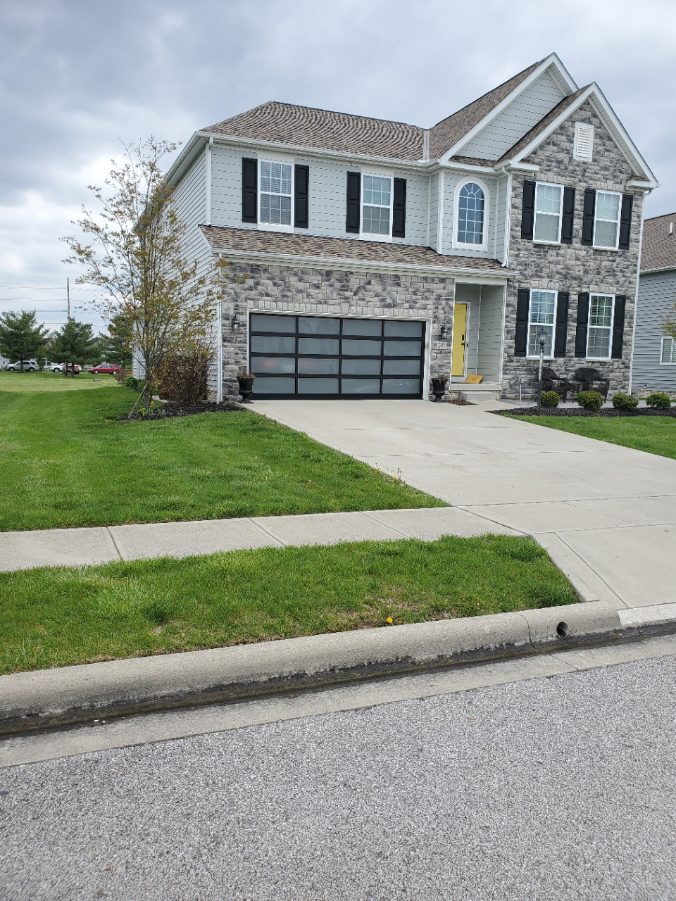 Suburban two-story stone house with a gray garage, green lawn, and driveway on a cloudy day