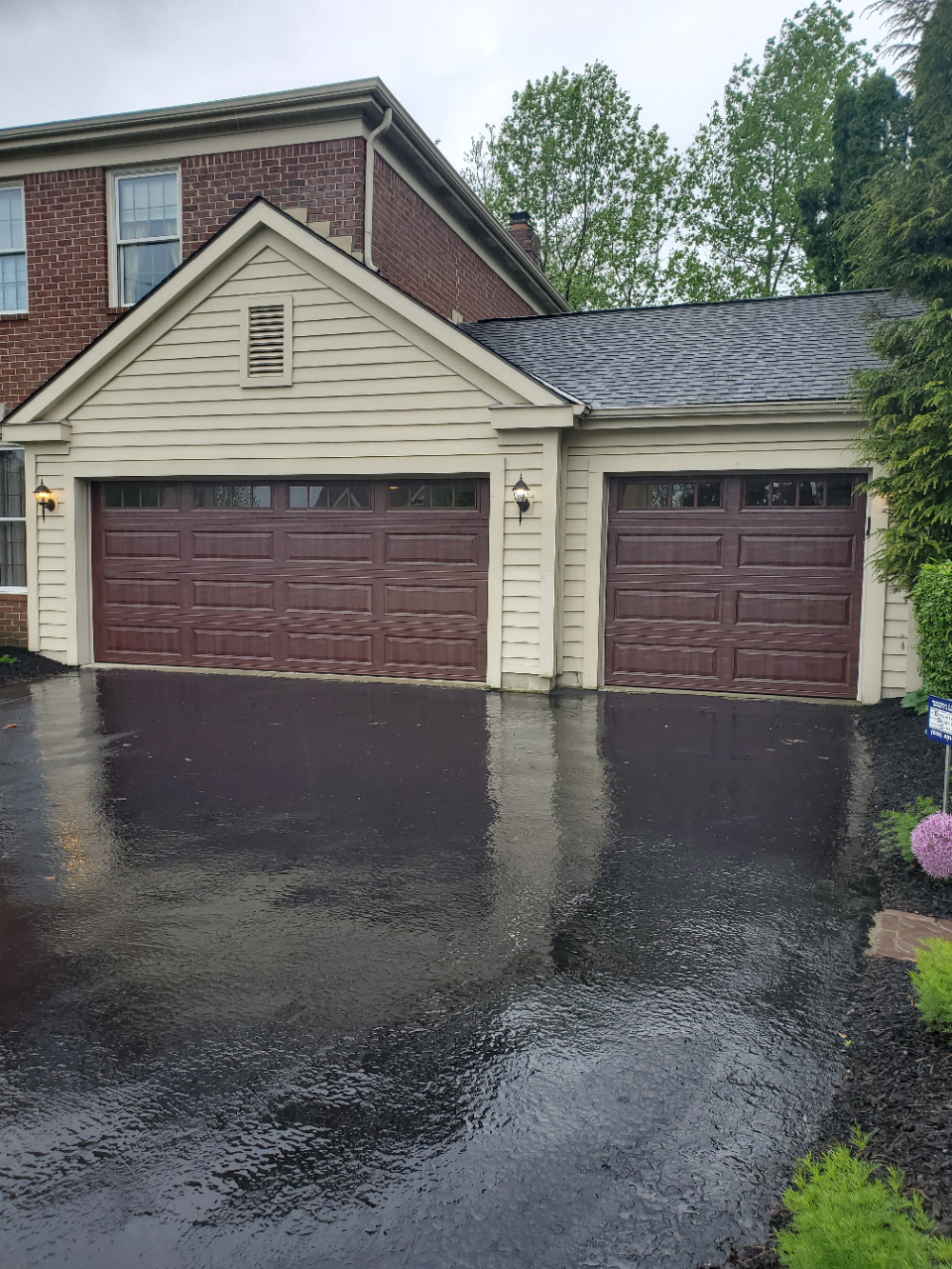 Two-car garage with dark wet driveway after rain and beige siding on a brick house