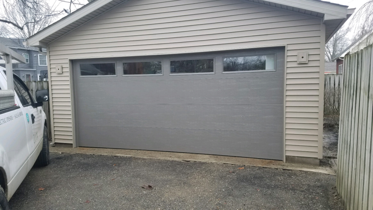Gray garage with a closed door and small windows, viewed from a driveway between a white truck and a fence.