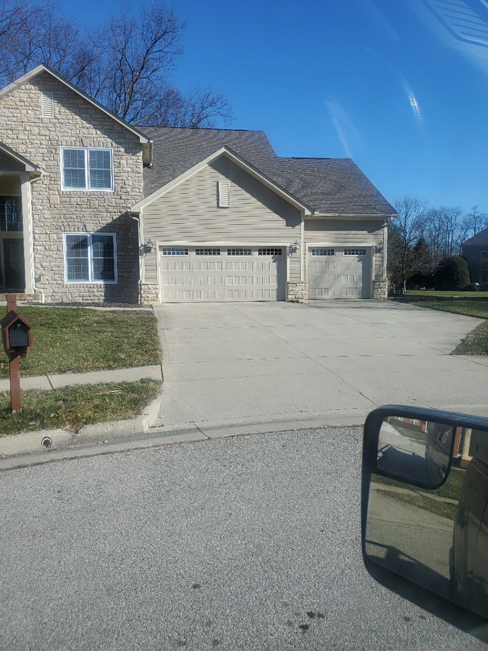 Suburban stone house with three-car garage and driveway under a clear blue sky; car side mirror visible.