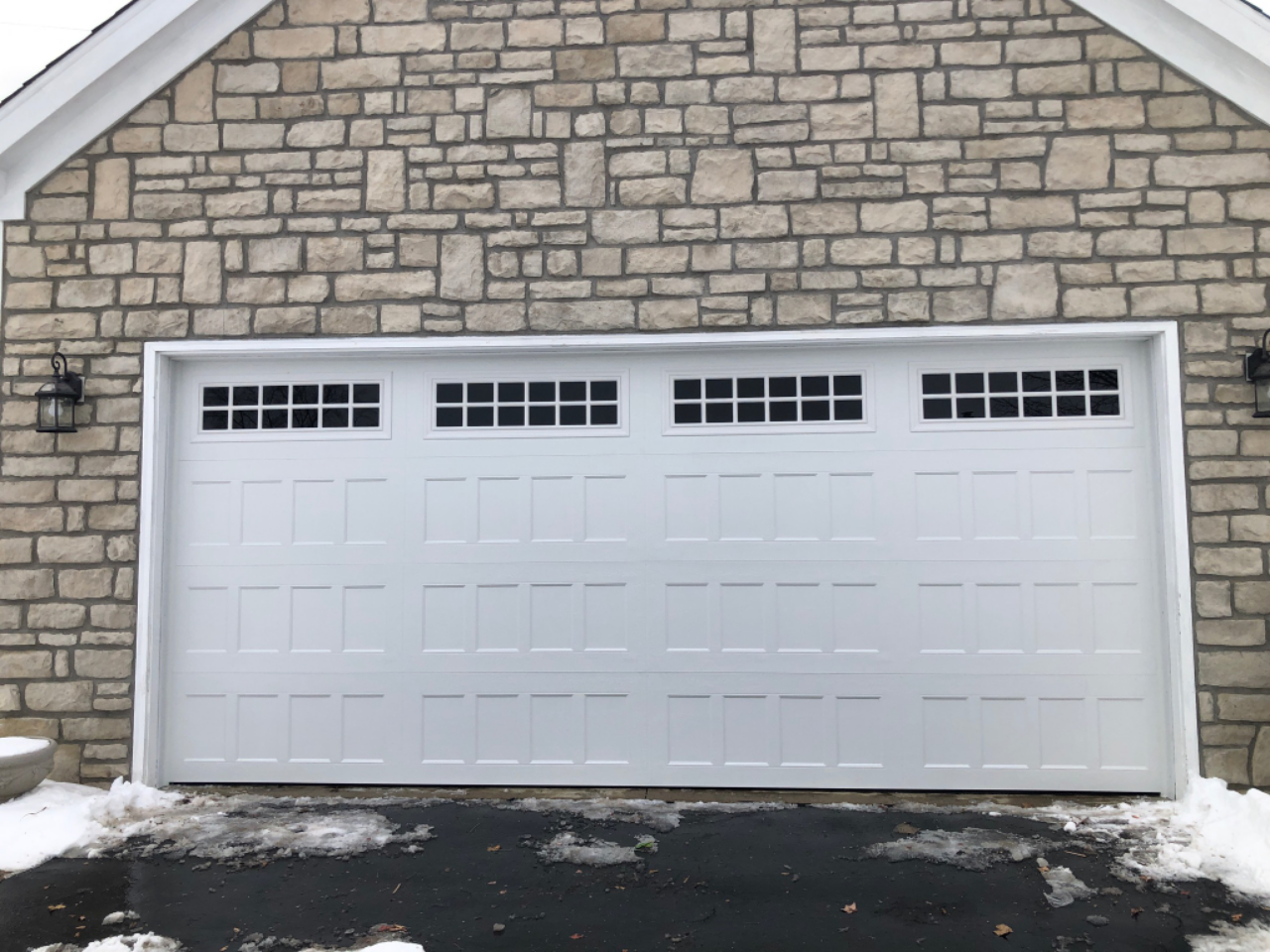 Snow-covered driveway in front of a beige stone house with a white garage door.