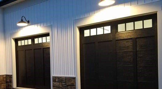 Two dark garage doors on a light-sided building, lit by exterior wall lamps at dusk