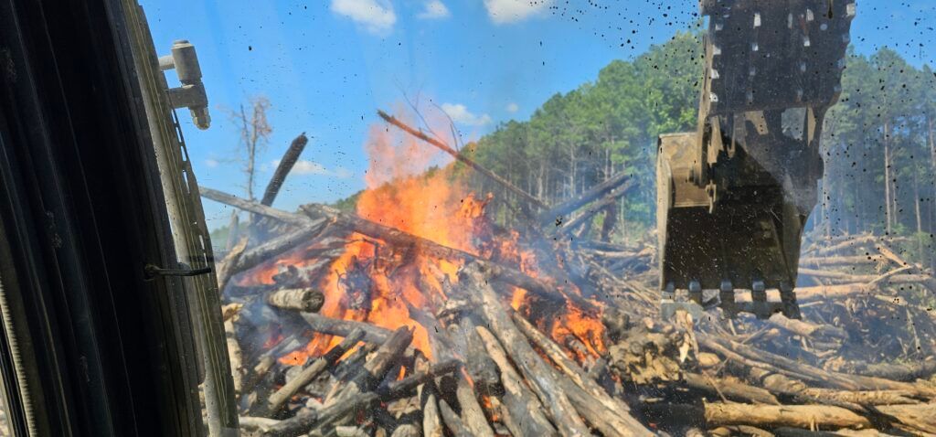 Large outdoor fire burning through debris near a blue corrugated wall and trees, with smoke and a hanging black bag