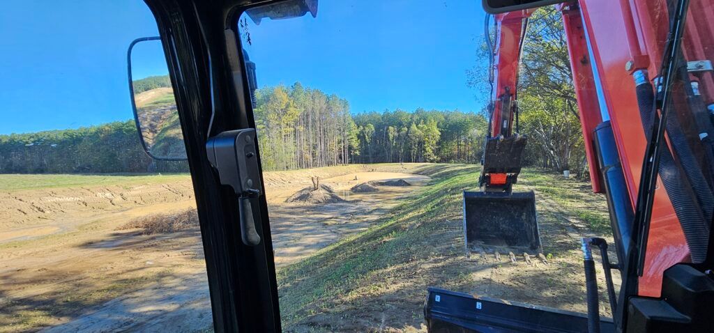 View from an excavator cab over a dirt field with an orange arm and bucket on the right, trees in the distance