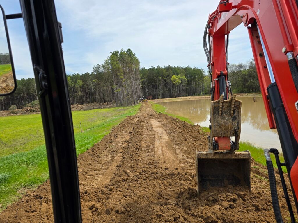 Excavator spreading dirt along a muddy path beside a grassy field and water